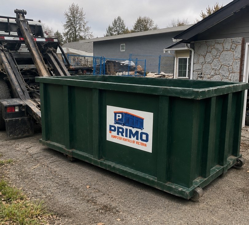 Green roll-off dumpster with the Primo decal, placed beside a house while the hauling truck pulls away